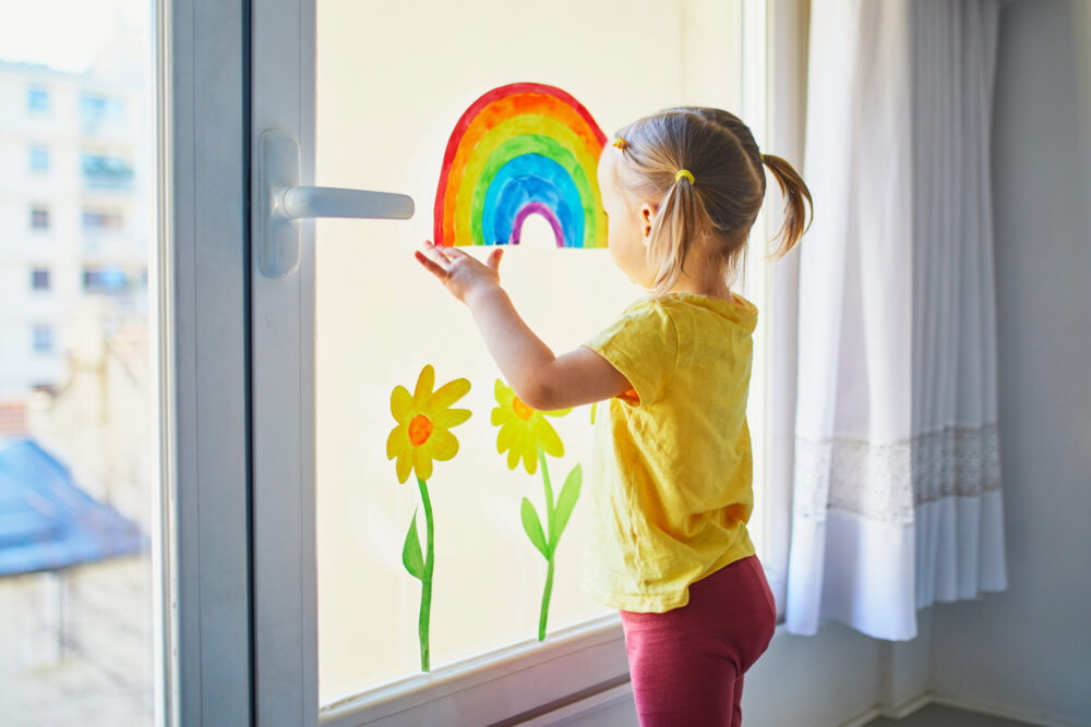 Adorable toddler girl attaching drawing of rainbow to window glass as sign of hope